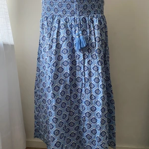 Blå mönstrad lång kjol från Gina Tricot - Säljer en blå mönstrad lång kjol från Gina Tricot med smockdetaljer upptill och dekorativa tofsar. Perfekt för en somrig look med sitt lätta och luftiga material. På ena snöret har några pärlor åkt av men de är inget man tänker på.🌸