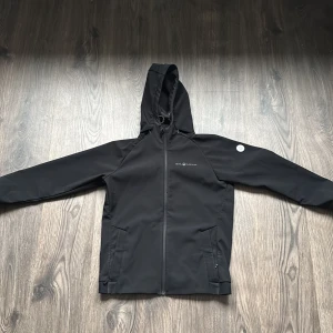 Svart softshelljacka med huva sail racing  - Snygg svart softshelljacka från Sail Racing med huva och dragkedja framtill. Jackan har långa ärmar och praktiska fickor med dragkedja. Perfekt för utomhusaktiviteter. PRIS KAN DISKUTERAS!