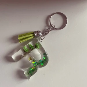 Resin Nyckelring💚 - Unik nyckelring med en transparent bokstav fylld med gröna glitterdetaljer och små blommor. Den har en grön tofs som ger en extra touch. Perfekt för att sätta personlig prägel på dina nycklar eller väskor.💚