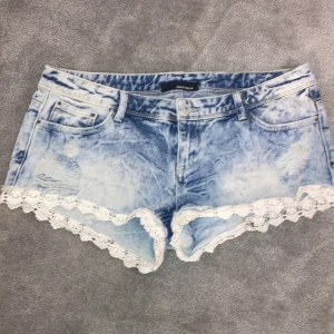 Blå jeansshorts med spets från Tally Weijl - Snygga ljusblå jeansshorts från Tally Weijl med coola slitningar och vit spetsdetalj längs kanten. Perfekta för en avslappnad sommarlook. De har en knapp och dragkedja framtill samt klassiska fem fickor. Mått tvärsöver:41 längde:22cm. Står inte storlek men passar mig som är S-M
