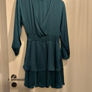 Mörkgrön klänning aldrig använd  från Zara - Knälång mörkgrön klänning från Zara med långa ärmar och smockdetalj i midjan. Perfekt för en stilren look med sitt flytande tyg och v-ringade design. Klänningen har en luftig känsla och passar bra till olika tillfällen.