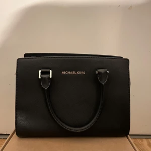 Svart handväska från Michael Kors - Elegant svart handväska från Michael Kors i skinn. Väskan har både handtag och en avtagbar axelrem för flexibilitet. Den har en stilren design med metallfötter på undersidan för extra skydd. Perfekt för att bära dina dagliga nödvändigheter med stil. Lite slitningar på texten och under men inget man tänker på❣️