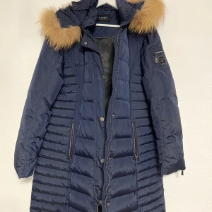 Jacka By Junge äkta päls  - Säljer en snygg mörkblå dunjacka från By Junge med avtagbar huva och äkta pälsdetalj. Jackan har långa ärmar, dragkedja och knappar framtill samt praktiska fickor med dragkedja. Finns i Åstorp.