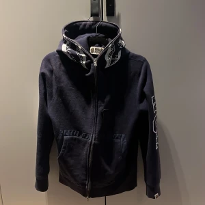 Mörkblå hoodie från A Bathing Ape - Säljer en cool mörkblå hoodie från A Bathing Ape med det ikoniska hajmönstret på huvan. Hoodien har en dragkedja framtill och praktiska fickor. Perfekt för streetwear-entusiaster!