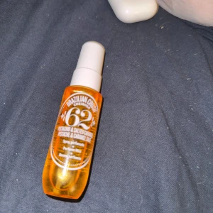 Brazilian Crush Cheirosa '62 - En doftspray från Sol de Janeiro med en varm och söt doft av pistage och saltad karamell. Flaskan är orange med en vit spraykork och har en stilren design. Perfekt för att ge en exotisk touch till din dag.