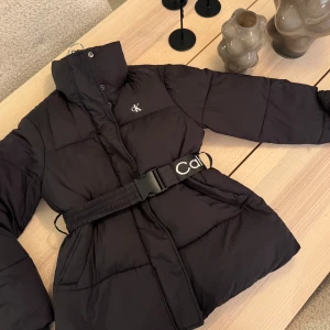Svart pufferjacka från Calvin Klein - Snygg svart pufferjacka från Calvin Klein med ett justerbart bälte i midjan. Jackan har en hög krage och stängs med dragkedja och knappar. Perfekt för kyliga dagar med sin vadderade design.