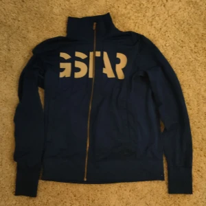 Blå zip från G-Star - Säljer en snygg blå jacka från G-Star med dragkedja och hög krage. Jackan har långa ärmar och ett stort G-Star-tryck på ryggen. Lite smutsig, väl behandlad, släng in den i tvätten så är den som ny. Kan snackas ner.