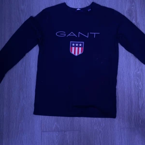 Svart tröja från Gant - Säljer en stilren svart tröja från Gant med deras ikoniska logga och sköld på bröstet. Tröjan har långa ärmar och en rund halsringning. Perfekt för en avslappnad och snygg look.