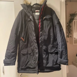 Svart parkajacka från Fjällräven - Säljer en svart parkajacka från Fjällräven med huva och pälsdetaljer. Jackan har dragkedja och knappar framtill samt flera praktiska fickor. Nypris 6000