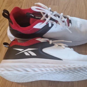 Vita och röda sneakers från Reebok - Snygga vita sneakers från Reebok med röda och svarta detaljer. Skorna har en bekväm sula och klassisk snörning. Perfekta för en sportig look eller vardagsbruk. Sula 23,3 cm