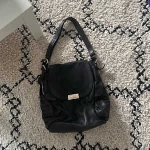 Svart axelväska i skinn - Snygg svart axelväska i skinn med praktisk dragkedja och en framficka med knapp. Perfekt för att bära med sig det nödvändigaste. Väskan har en justerbar axelrem och stilren design.