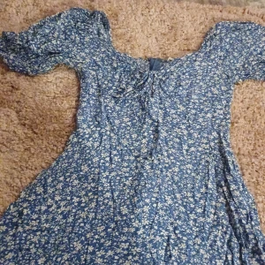 Blå blommig klänning - Charmig blå klänning med ett fint blommigt mönster i vitt. Klänningen har korta ärmar och en söt knytning framtill. Perfekt för en somrig look! Dragkedja på ryggen