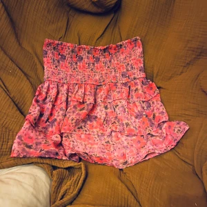 Rosa blommig kjol med volanger - Charmig rosa kjol med blommigt mönster och volanger. Den har en elastisk midja för bekväm passform och är perfekt för en somrig look.