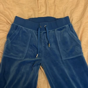 Blå mjukisbyxor från Juicy Couture - Snygga blå mjukisbyxor från Juicy Couture i mjukt material. Byxorna har snörning i midjan och praktiska fickor med knappar bak. Perfekta för en avslappnad stil med en touch av lyx.