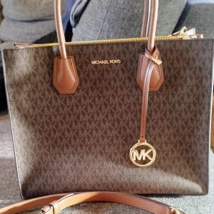 Brun handväska från Michael Kors - Snygg brun handväska från Michael Kors med det klassiska MK-mönstret. Väskan har två handtag och en dragkedja upptill. Perfekt för att bära dina dagliga nödvändigheter med stil.