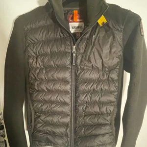 Svart jacka från Parajumpers - Snygg svart/mörk grön jacka från Parajumpers med quiltad design och dragkedja framtill. Jackan har en cool patch på ärmen och är perfekt för kyligare dagar. Den kombinerar stil och funktionalitet med sitt unika materialval. Jackans nypris ligger på 4000 och skicket är 9/10. Kvitto och extra knappar och prislappar medföljer.