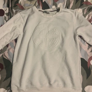 Vit tröja från Stone Island - Säljer en stilren vit tröja från Stone Island med deras ikoniska logga broderad på bröstet. Tröjan har långa ärmar och en klassisk passform. Perfekt för en avslappnad och trendig look.