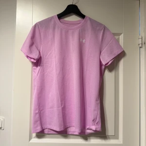 Ljusrosa t-shirt från Under Armour - Säljer en ljusrosa t-shirt från Under Armour. Den har en enkel design med korta ärmar och en rund halsringning. Perfekt för träning eller vardagsbruk. Under Armour-loggan syns diskret på bröstet.