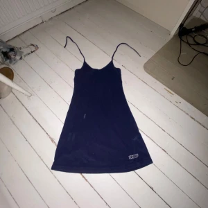 Mörkblå klänning från BikBok - Säljer en mörkblå klänning från BikBok med smala axelband som knyts i nacken samt ett litet tryck på framsidan vid låret och på ryggen. Perfekt för en picknick på stranden om du blir bjuden på don’t härligt 🤷‍♂️. Klänningen har en enkel design och är tillverkad i ett handduks liknande material.