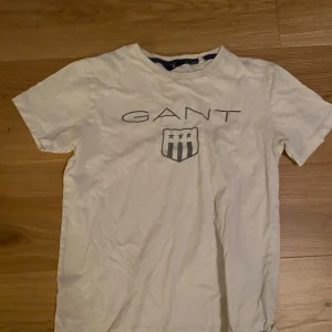 Vit t-shirt från Gant - Säljer en vit t-shirt från Gant med tryckt logotyp och emblem på framsidan. T-shirten har en klassisk rund hals och korta ärmar. Perfekt för en avslappnad stil.