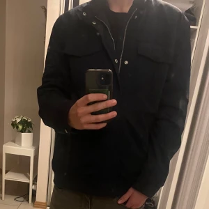 Mörkblå fieldjacka från Hampton Republic - Snygg mörkblå jacka från Hampton Republic med krage och knappar. Jackan har två bröstfickor och långa ärmar med knappdetaljer vid ärmsluten. Perfekt för en stilren look.