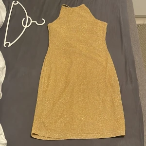 Guldglittrig klänning - Snygg guldglittrig klänning med öppen rygg och knytband. Den är armlös och man kan knyta till den thightare där bak. Lite kort men super snygg och glittrig. Köpt på H&M för några år sedan. Aldrig använt, perfekt till utgångar 
