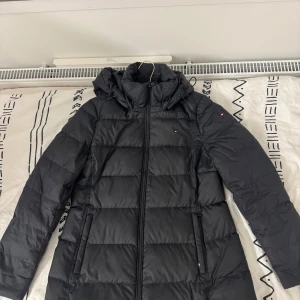 Svart dunjacka från Tommy Hilfiger - Snygg svart dunjacka från Tommy Hilfiger med dragkedja och hög krage. Jackan har långa ärmar och är perfekt för kyliga dagar. Den quiltade designen ger en stilren look.