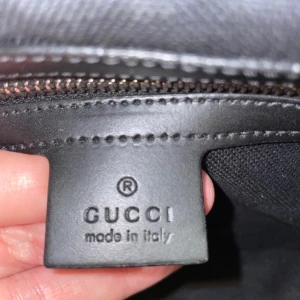 Svart axelväska från Gucci - Snygg svart axelväska från Gucci med klassiskt GG-mönster. Väskan har en justerbar axelrem i rött och blått samt en praktisk dragkedja framtill. Perfekt för att bära dina viktigaste saker med stil.