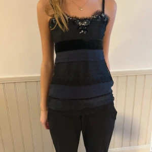 Svart och blå festtopp med pärlor - Snygg svart och blå linnetopp med smala axelband och dekorativa pärlor vid halsringningen. Toppen har en randig design med olika texturer och en knappar i ryggen.