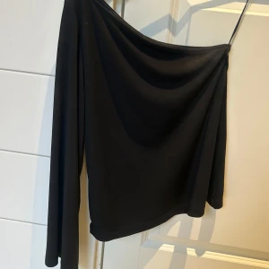 Svart oneshoulder topp från Lindex - Snygg svart oneshoulder topp från Lindex med en lång ärm. Perfekt för en stilren look. Tillverkad i ett mjukt och bekvämt material. Endast använd en gång så är i väldigt fint skick. 