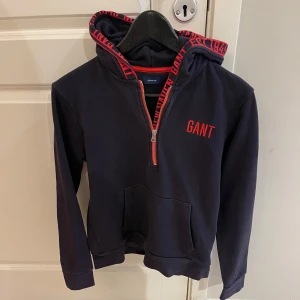 Svart hoodie från GANT - Säljer en snygg svart hoodie från GANT i mycket bra skick. Den har en halv dragkedja och röd text på bröstet samt längs huvan. Perfekt för höst och vår, med en skön passform och känguruficka framtill.