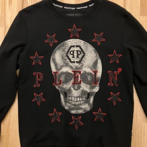 Svart tröja med dödskallemotiv från Philipp Plein - Snygg svart tröja från Philipp Plein med ett stort dödskallemotiv och röda stjärnor på framsidan. Tröjan har långa ärmar och en rund halsringning med märkets logotyp på baksidan. Perfekt för en edgy look!