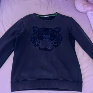 Svart sweatshirt från Kenzo - Snygg svart sweatshirt från Kenzo med ett coolt tigerbroderi på framsidan. Tröjan har långa ärmar och ribbade muddar vid ärmslut och nederkant. Perfekt för en stilren look!