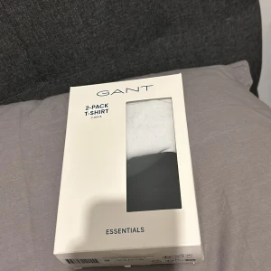 2-pack T-shirt från GANT - Säljer ett 2-pack T-shirts från GANT i storlek 170 cm, perfekt för tonåringar. Ena t-shirten är svart och den andra är vit, båda med rund hals och GANT-logga på bröstet. De är i nytt skick och endast använda en gång. Kommer i originalförpackning. 