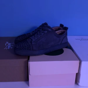 Grå sneakers med nitar från Christian Louboutin - Snygga grå  sneakers från Christian Louboutin med nitar på tån. Skorna har en klassisk snörning och en röd sula som ger en ikonisk touch. Perfekta för att ge din stil en edgy känsla.