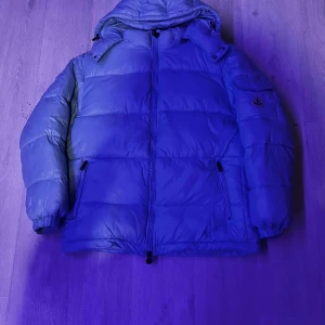 Blå dunjacka från Moncler - Säljer en snygg blå dunjacka från Moncler med huva och dragkedja. Jackan har en pufferdesign och långa ärmar, perfekt för kyliga dagar. Den är både stilren och funktionell.