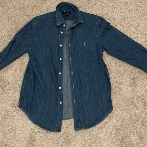Blå jeansskjorta från Polo Ralph Lauren - Säljer en snygg blå jeansskjorta från Polo Ralph Lauren med klassisk krage och knappar framtill. Skjortan har ett broderat emblem på bröstet och långa ärmar. Perfekt för en stilren look!
