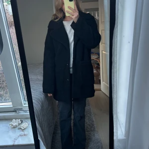 Svart kappa - Snygg svart kappa i teddy material. Den har långa ärmar och en stilren look som passar perfekt för kyligare dagar. Perfekt för att hålla stilen i höst och vinter.