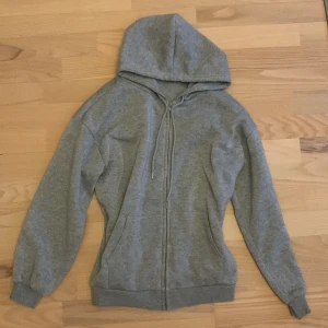 Grå hoodie med dragkedja - Säljer en mysig grå hoodie i bra skick. Den har en dragkedja framtill och en praktisk huva. Perfekt för kyliga dagar eller som ett extra lager. Passar till det mesta och är superbekväm!