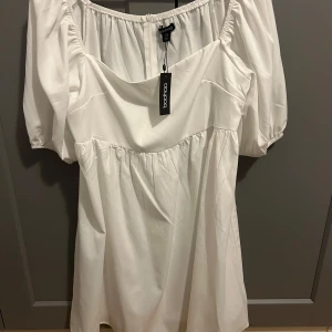 Vit klänning från Boohoo - Säljer en stilren vit klänning från Boohoo med korta puffärmar. Klänningen har en luftig passform. Perfekt för till exempel studenten. Helt oanvänd med prislapp kvar.