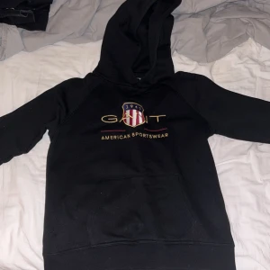Svart hoodie från GANT - Säljer en svart hoodie från GANT med klassisk logga och texten 'American Sportswear' på bröstet. Tröjan har en bekväm huva och en stor ficka framtill. Perfekt för en avslappnad stil. Äkta storlek 16 years/ 167cm