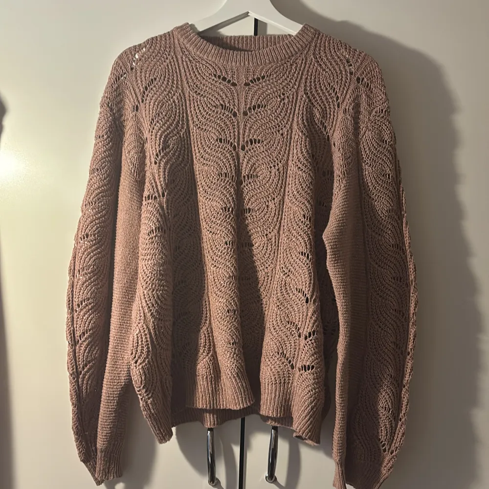 Säljer en mysig rosa stickad tröja med ett vackert hålmönster. Tröjan har långa ärmar i ballongstil och en rund halsringning.. Neuletakit & Villapaidat.