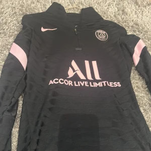 Svart långärmad PSG-tröja - Säljer en snygg svart långärmad PSG-tröja från Nike. Den har rosa detaljer och texten 'Accor Live Limitless' på framsidan. Tröjan har också 'Visit Rwanda' på baksidan och PSG-loggan på bröstet. Perfekt för fans av Paris Saint-Germain!