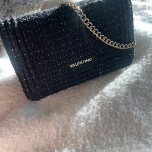 Svart axelväska från Valentino - Elegant svart axelväska från Valentino med en stilren kedjerem. Väskan har en glittrig yta och ett diskret mönster. Perfekt för att ge en touch av glamour till din outfit eller en julklapp/present till dig eller någon du tycker om.