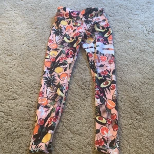 Färgglada leggings från Stronger - Säljer ett par supercoola leggings från Stronger i storlek S. De har ett färgglatt fruktmönster med avokado, apelsiner och körsbär på en rosa bakgrund. Tillverkade av 85% återvunnen polyester och 15% elastan, vilket gör dem både miljövänliga och stretchiga. Perfekta för träning eller en färgglad vardagslook!