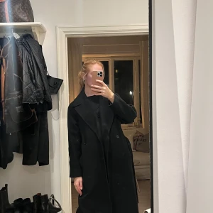 Svart ullkappa från Weekday - Säljer en  svart kappa från Weekday i mycket bra skick. Den har en klassisk design med knappar och ett bälte i midjan. Supervarm & skön då det är ull i. Storlek xs men lite obersized skulle jag säga. Några små noppror på. Köpt för 1800kr