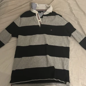 Randig pikétröja från Tommy Hilfiger - Säljer en snygg randig pikétröja från Tommy Hilfiger i grått och svart. Tröjan har en klassisk krage med knappar och är perfekt för en avslappnad stil. Den är i bra skick och passar både till vardags och lite finare tillfällen.