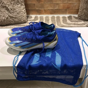 ADIDAS X CRAZYFAST MESSI ELITE FG SPARK GEN10S - LUCID BLUE/BLUE BURST/VIT - Snygga blå fotbollsskor från Adidas med vita och guldiga detaljer. Skorna har snörning och en slitstark sula med dobbar för optimalt grepp på både gräs och konstgräs planen. Perfekta för fotbollsträning och matcher. Skorna är i helt ny skick har bara haft på de 2-3 gånger, skorna är messis egna speciella skor och är även helt äkta köpt för 3000kr  