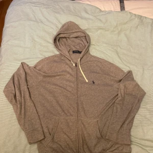 Grå hoodie från Ralph Lauren - Säljer en stilren grå hoodie från Ralph Lauren med dragkedja och justerbar huva. Perfekt för en avslappnad look. Den har långa ärmar och en liten logga på bröstet. Skön och enkel att matcha med andra plagg.