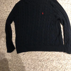 Marinblå stickad tröja från Ralph Lauren - Säljer en stilren svart stickad tröja från Ralph Lauren med det klassiska röda logotypen på bröstet. Tröjan har ett flätmönster och långa ärmar, perfekt för kyligare dagar Storlek L men lite stor i storleken, priset är inte hugget i stenen. Öppen för byten 